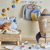 Cosmina Rocket Seagrass Basket with Lid in natural woven seagrass, shaped like a rocket with a black fin, displayed in a nursery beside toys and a wall mobile.