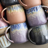 Close-up of Cloe stoneware mugs with C-shaped handles, each glazed in a different colourway with drip streaks from the rim over a speckled lower body, stacked together.