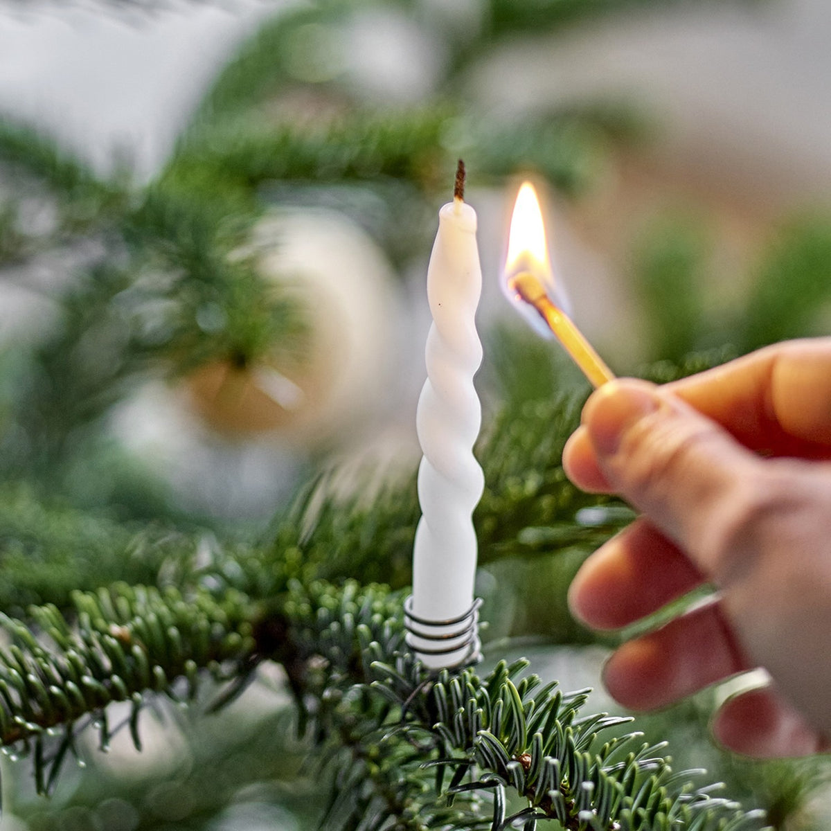 A hand lights a slim white spiral Twist Candle in a holder on a Christmas tree branch, with baubles blurred in the background.