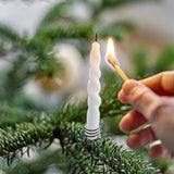 A hand lights a slim white spiral Twist Candle in a holder on a Christmas tree branch, with baubles blurred in the background.