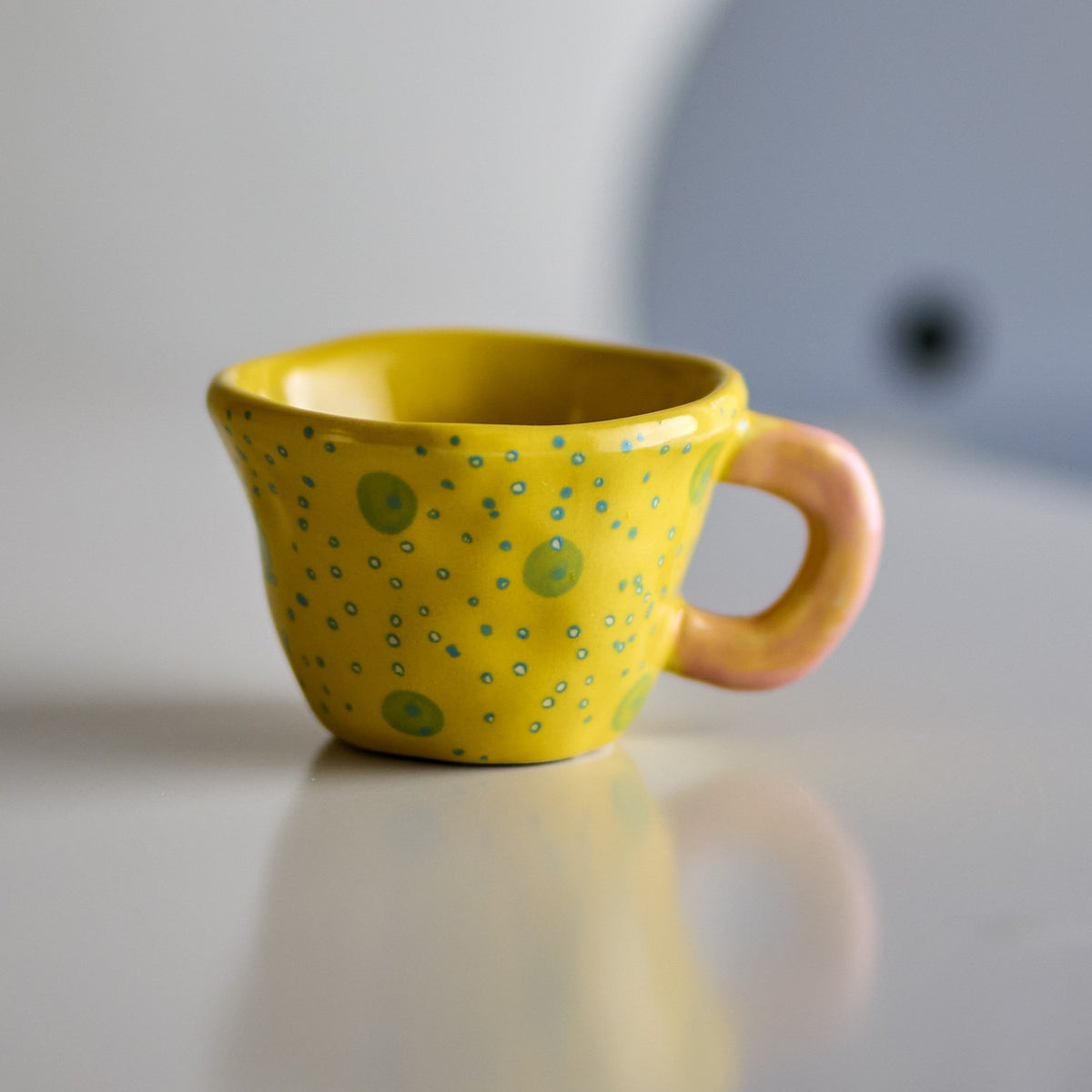Nini Stoneware Cup 100ml in Yellow, a small handmade-style ceramic cup with a pink loop handle and hand-painted spot and dot pattern, shown against a soft grey background.