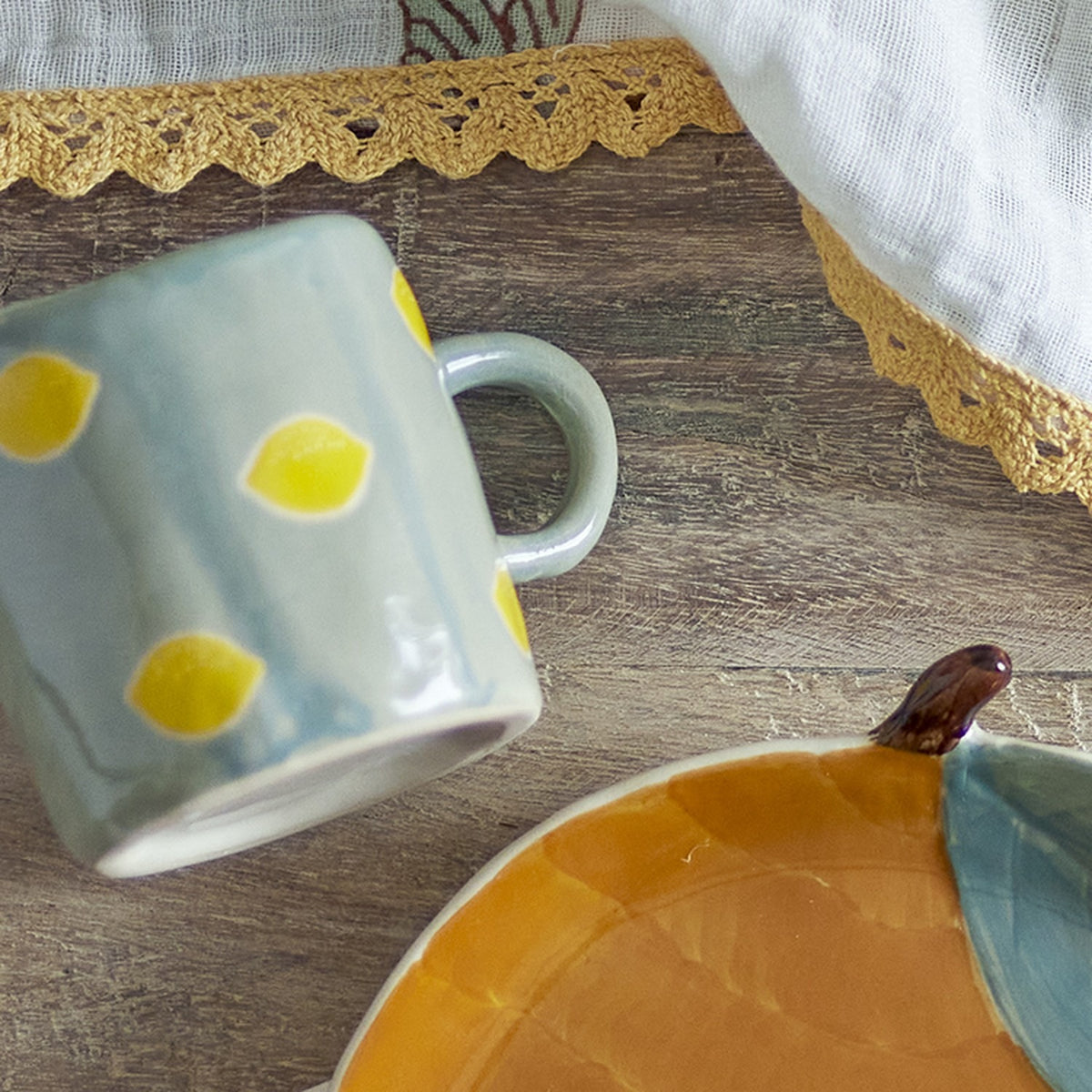 Agnes Stoneware Cup 230ml in green with yellow lemon dots and a curved handle, glossy ceramic finish, shown on a wood surface beside a mustard-glazed plate.