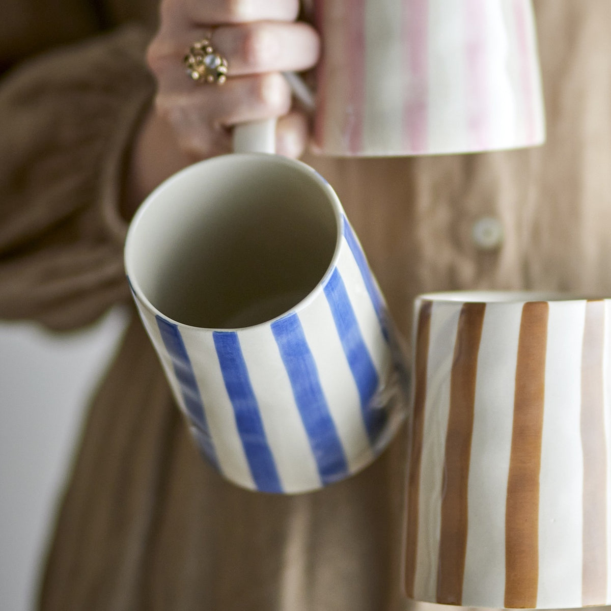 Begonia Striped Stoneware Mug 440ml in blue, held by hand, showing the handle and interior, with pink and brown striped mugs visible in the background.