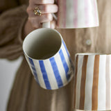 Begonia Striped Stoneware Mug 440ml in blue, held by hand, showing the handle and interior, with pink and brown striped mugs visible in the background.