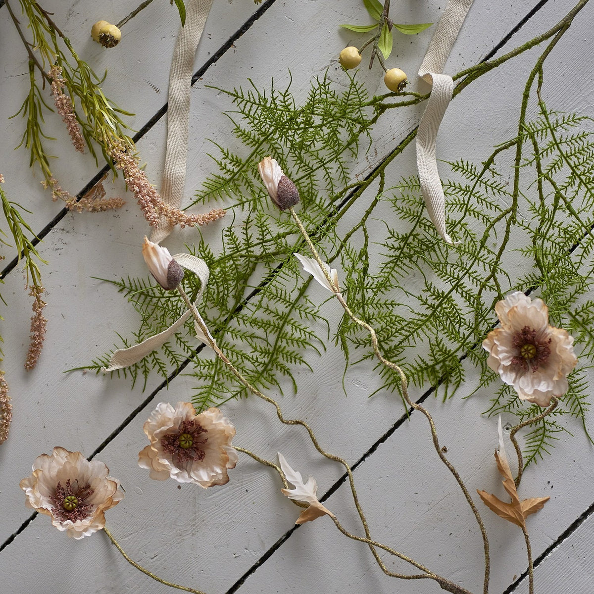 Poppy Artificial Stem in beige with ruffled open flower heads and closed buds on twisting stems, styled with fern fronds and ribbon on a light wood-slat surface.