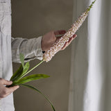 Hand holding the Foxtail Artificial Stem 73cm in pink, showing the tapered flower spike and long slender green leaves against a neutral curtained background.