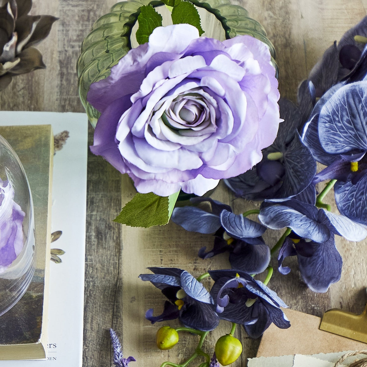 Artificial purple rose stem with a ruffled open bloom and green serrated leaves, displayed in a glass vase among blue iris stems on a wooden surface.