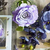 Artificial purple rose stem with a ruffled open bloom and green serrated leaves, displayed in a glass vase among blue iris stems on a wooden surface.