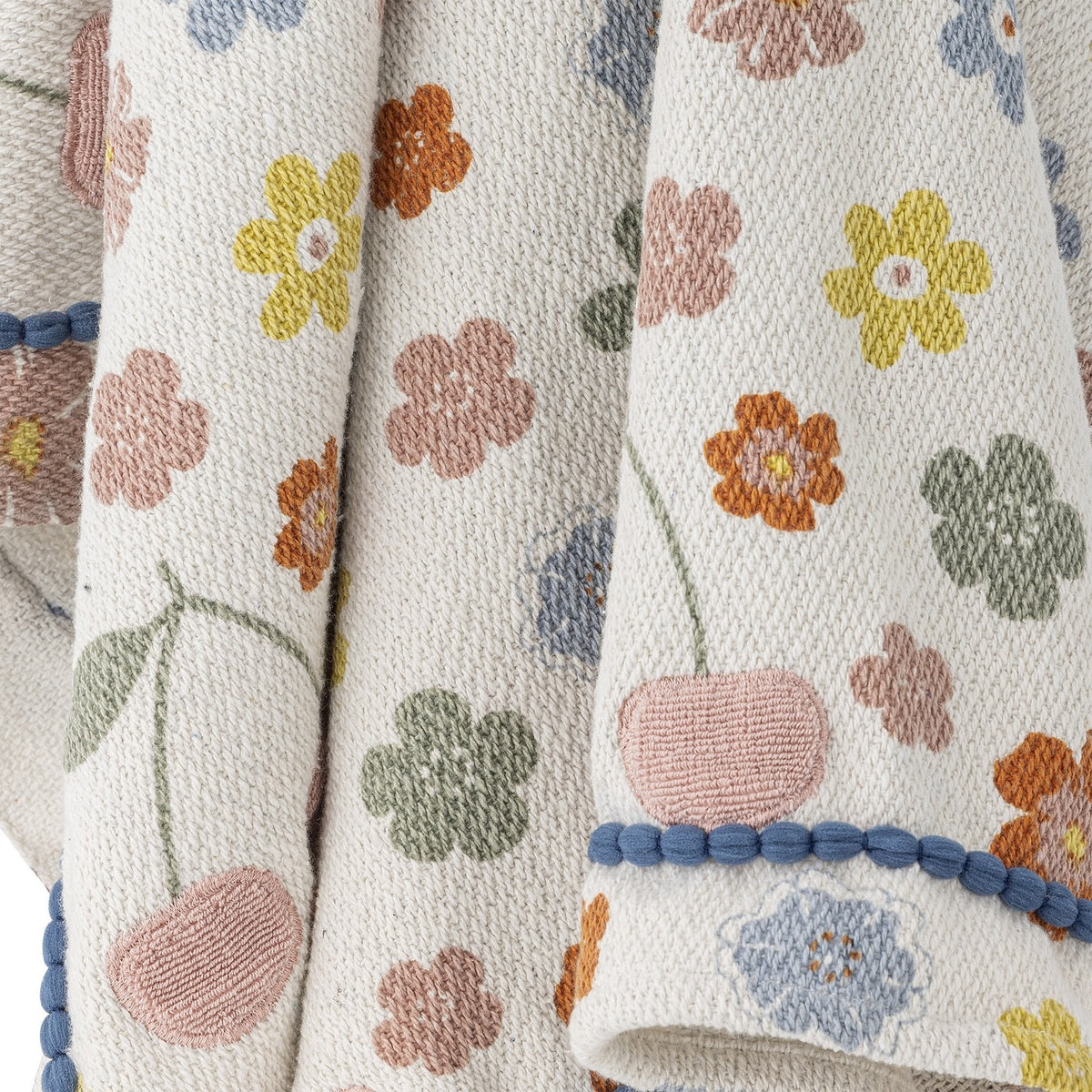 Cherry Floral Cotton Throw in Multi, close-up of textured woven fabric with pastel flowers and embroidered cherries, finished with a blue pom-pom trim edge.