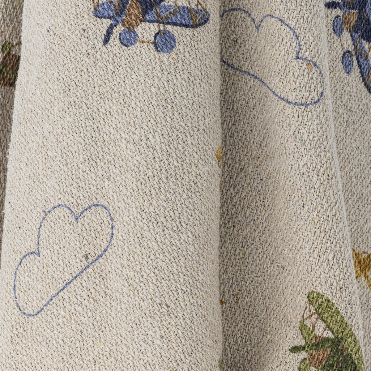 Plane Cotton Throw in beige, close-up of folded cotton fabric showing outlined cloud motifs and a glimpse of a biplane print.