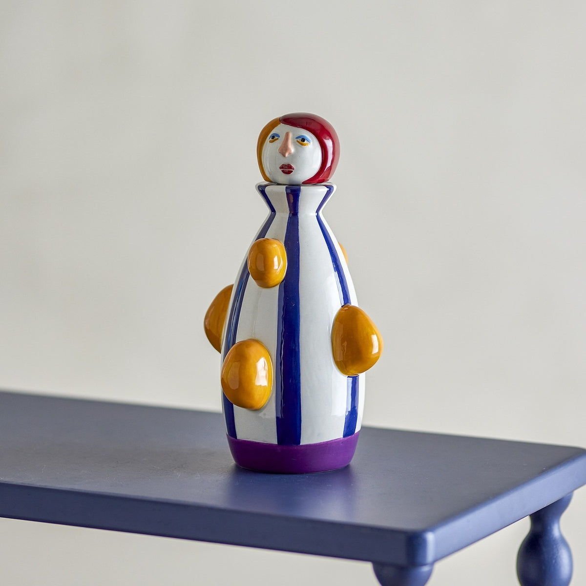 Conneli Ceramic Candle Holder in Multi, a bottle-shaped dolomite figure with a painted face, red bobbed hair, blue stripes, orange raised bumps and a purple foot band, displayed on a blue table.