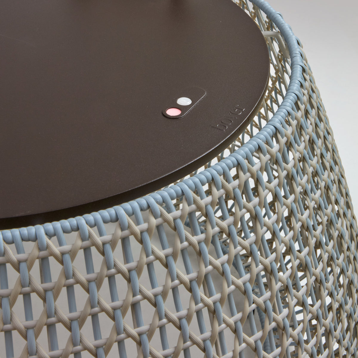 Close-up of the Nans Bag/38 outdoor floor lamp showing its dark round top with control buttons and brand marking, set above a woven shade in beige and light blue tones.