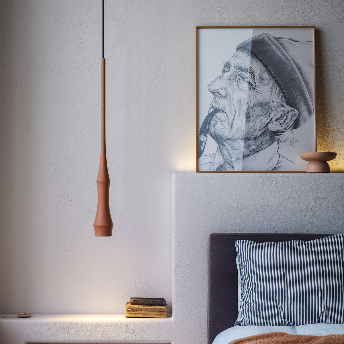 Slend 01 aluminium pendant light in terracotta, slim tapering form hanging beside a framed portrait sketch in a bedroom with striped pillows.