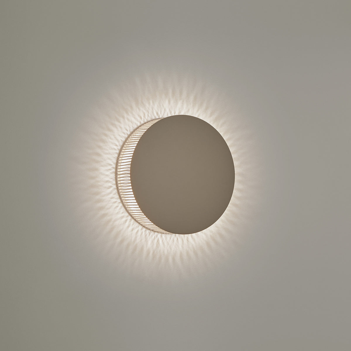 Helios A/01 round LED wall light with a solid oak brown disc front and beige cotton thread edge, casting a soft radiating halo on a plain light grey wall.
