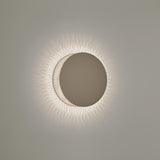 Helios A/01 round LED wall light with a solid oak brown disc front and beige cotton thread edge, casting a soft radiating halo on a plain light grey wall.