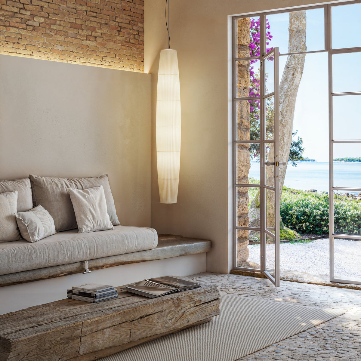 Maxi S/01 Ribbon LED pendant light with tall tapered white translucent pleated shade, hanging in a neutral living room beside glazed doors overlooking the sea.