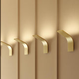 App Aluminium LED Wall Light in brushed brass, curved hook-shaped fixture mounted on panelled wall, shown in a row casting warm ambient uplight.