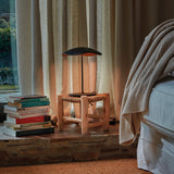 Valentina M/29 Table Lamp with wavy black and copper leaf-shaped shade on a slim black stem, shown on a wooden stool beside a bed with curtains and stacked books.