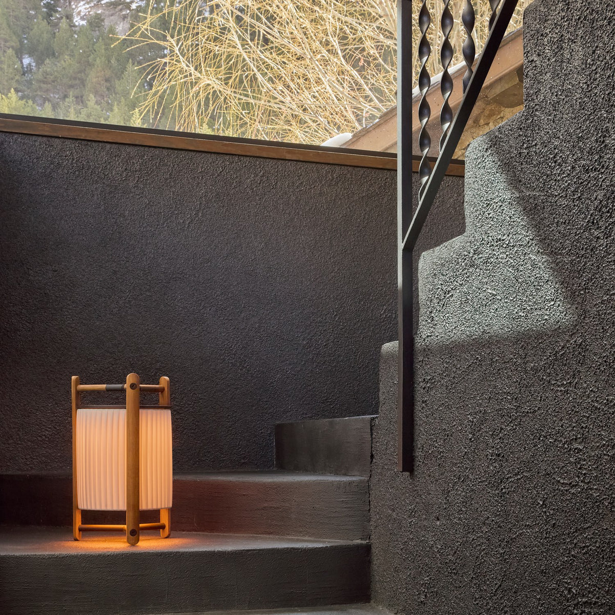 Kando M/40 Ribbon LED table lamp with a pleated white translucent cylinder shade in a wood-toned three-legged frame, lit outdoors on a dark stone step.