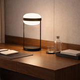 Beaded T Glass LED Table Lamp with an opal white flattened spherical shade, dark centre band and slim rods on a round black base, shown lit on a wooden desk.