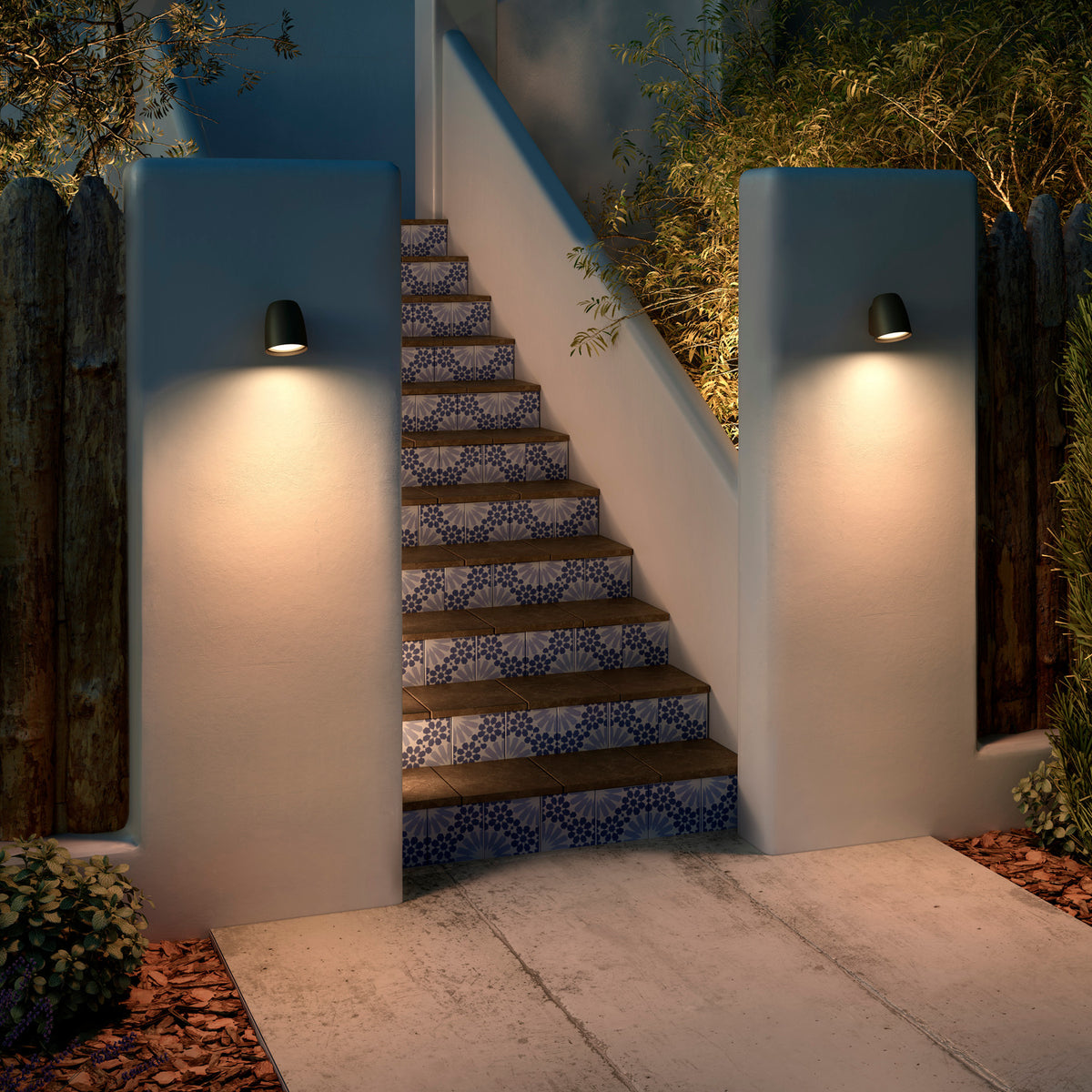 Nut A/01 outdoor wall lights with bell-shaped black heads mounted on rendered pillars, casting warm downward light onto a tiled staircase at dusk.