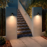Nut A/01 outdoor wall lights with bell-shaped black heads mounted on rendered pillars, casting warm downward light onto a tiled staircase at dusk.