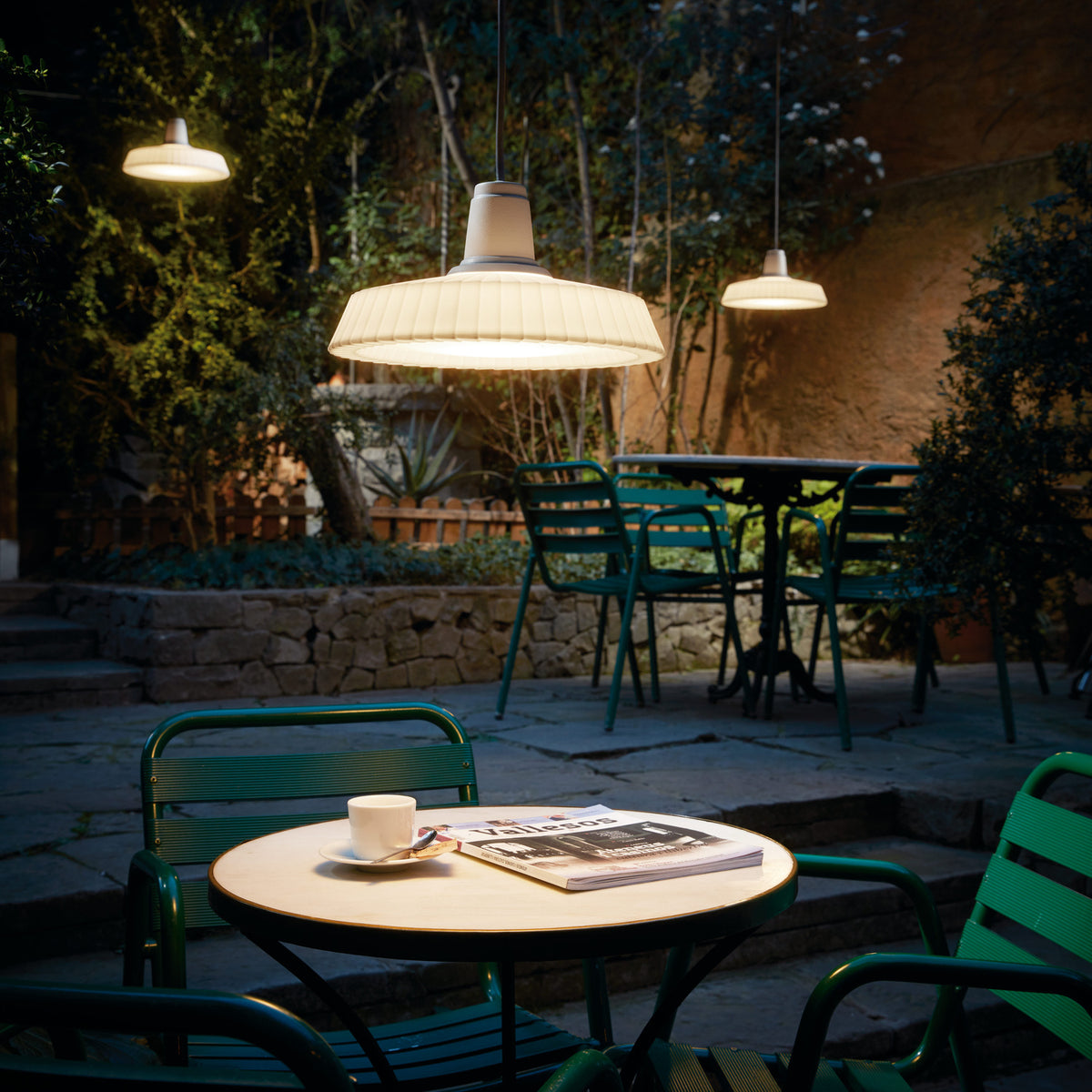 Marietta S/32 Dome LED Outdoor Pendant Light in white, with a pleated dome shade on a black cable, shown illuminated above a courtyard café table with green chairs at night.