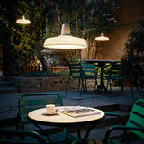 Marietta S/32 Dome LED Outdoor Pendant Light in white, with a pleated dome shade on a black cable, shown illuminated above a courtyard café table with green chairs at night.