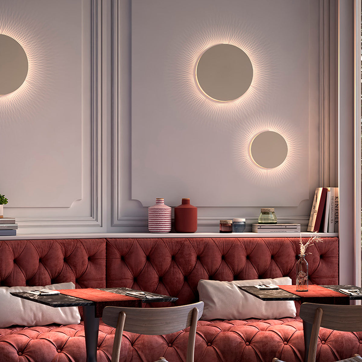 Round Helios A/02 LED wall lights with beige disc faces and warm fringed glow, mounted above a red tufted banquette in a dining setting.