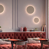 Round Helios A/02 LED wall lights with beige disc faces and warm fringed glow, mounted above a red tufted banquette in a dining setting.