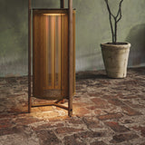 Kando P/111/R rechargeable outdoor floor lamp with sand cord cylinder, teak wood frame and three-legged base, glowing softly on a stone patio beside a potted plant.