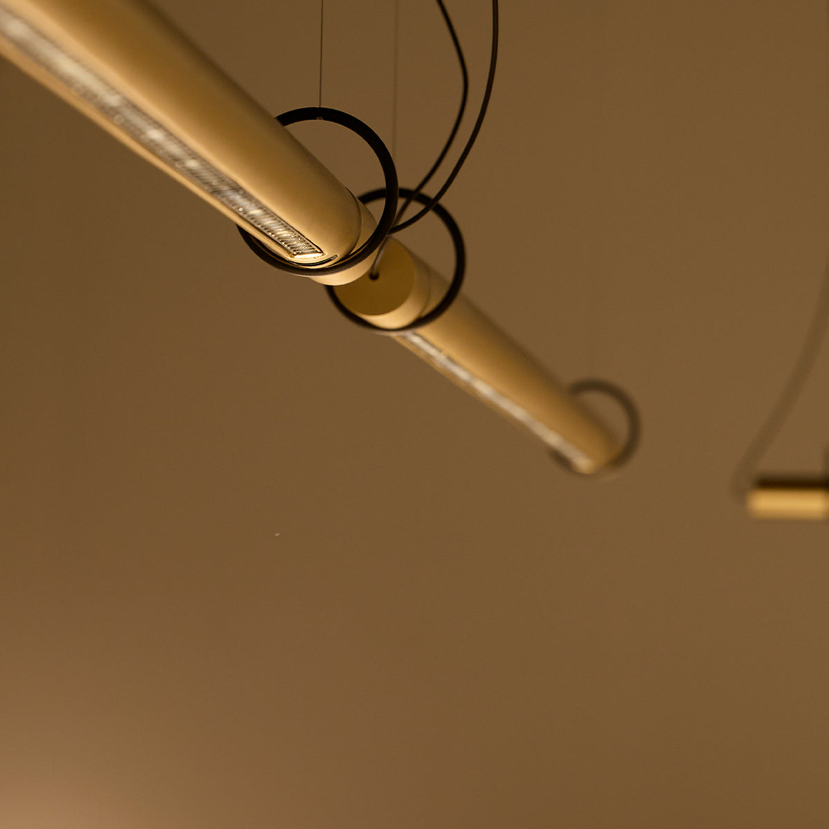 Close-up of the Solid SH Sens 100 Tube LED Pendant Light in brass-toned aluminium, showing its slim tube profile on fine suspension wires against a neutral background.