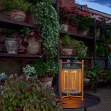 Kando M/40 Wood LED table lamp with cylindrical sand-coloured strand shade and teak wood frame, glowing warmly on an outdoor table among potted plants at dusk.