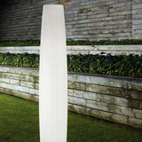 Maxi P/180 Cylinder LED Outdoor Floor Lamp in white polyethylene, a tall ribbed column fixture glowing softly on a grass lawn against a wooden fence and green hedges.
