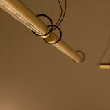 Close-up of the Solid SH Sens brass cylinder pendant light, a slim tube on fine wires with a line of light along its underside, shown at an angle against a warm-toned background.