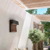 Sisal A/01 LED outdoor wall light with woven brown fibre front panel and angular hood, mounted on a rendered wall beside a pergola and garden setting.