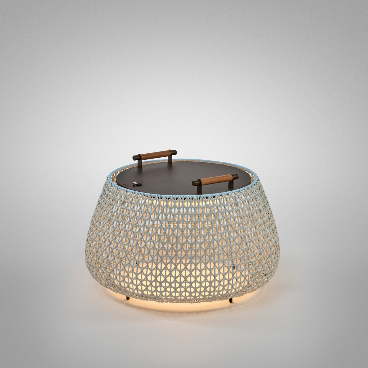 Nans Bag/38/R rechargeable outdoor floor lamp, a round woven basket shade in beige with light blue tint, dark lid and two wood handles, glowing LED light, on a plain light grey background.