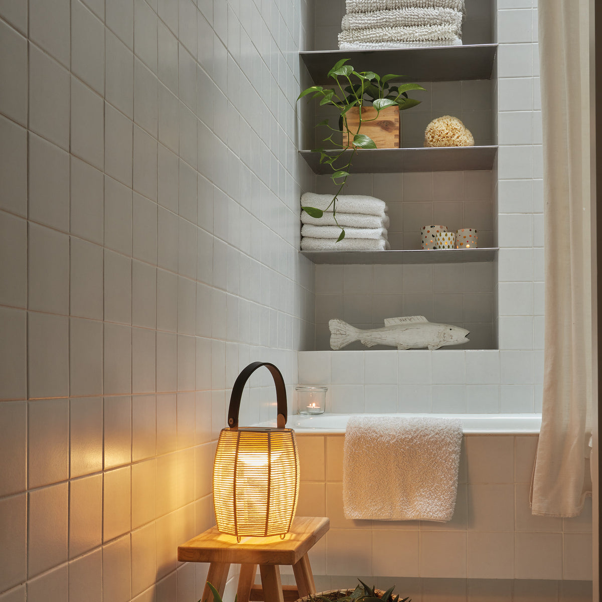 Tanit M/29/R rechargeable table lamp with brown woven fabric shade and curved handle, lit on a wooden stool beside a bathtub.