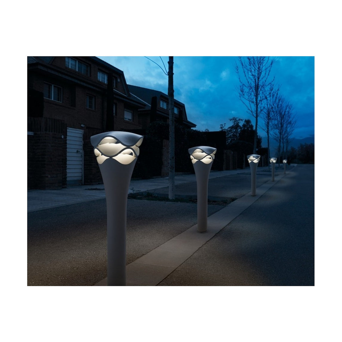 Cornet B/77 LED bollard lights in stone grey line a garden path at dusk, their slim flared stems topped with tiered wavy heads glowing warm white.
