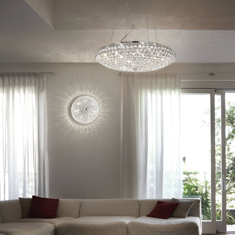 A modern living room features the Queen 9 Light Crystal Ceiling & Wall Light in chrome, casting intricate patterns across the walls. Below, a white sectional sofa adorned with red pillows is situated next to tall windows with sheer curtains, offering a view of the lush greenery outside.