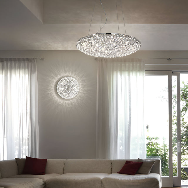 A modern living room features the Queen 9 Light Crystal Ceiling & Wall Light in chrome, casting intricate patterns across the walls. Below, a white sectional sofa adorned with red pillows is situated next to tall windows with sheer curtains, offering a view of the lush greenery outside.