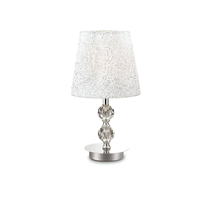 This elegant room decor piece, the Lumina Medium Crystal Table Lamp - Chrome, boasts a white fabric lampshade with a floral pattern set atop an ornate clear glass base. Ideal for modern classic style enthusiasts, it sits gracefully on a round metal platform against a plain white background.