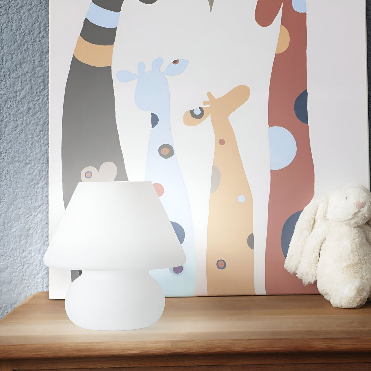 A Prato Large Table Lamp in white blown glass rests elegantly on a wooden dresser next to a plush white rabbit. In the background, a vibrant painting showcases five tall, abstract giraffes adorned with polka dots. The modern and elegant ambiance is enhanced by the wall's subtle blue-gray tone.