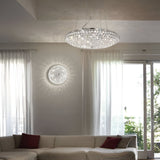 A modern living room boasts the Queen 5 Light Crystal Ceiling & Wall Light in chrome, with its versatile design creating intricate patterns on the ceiling and walls. Below, a beige sectional sofa complemented by red cushions is set beneath large windows draped with sheer white curtains, establishing a cozy yet elegant atmosphere.