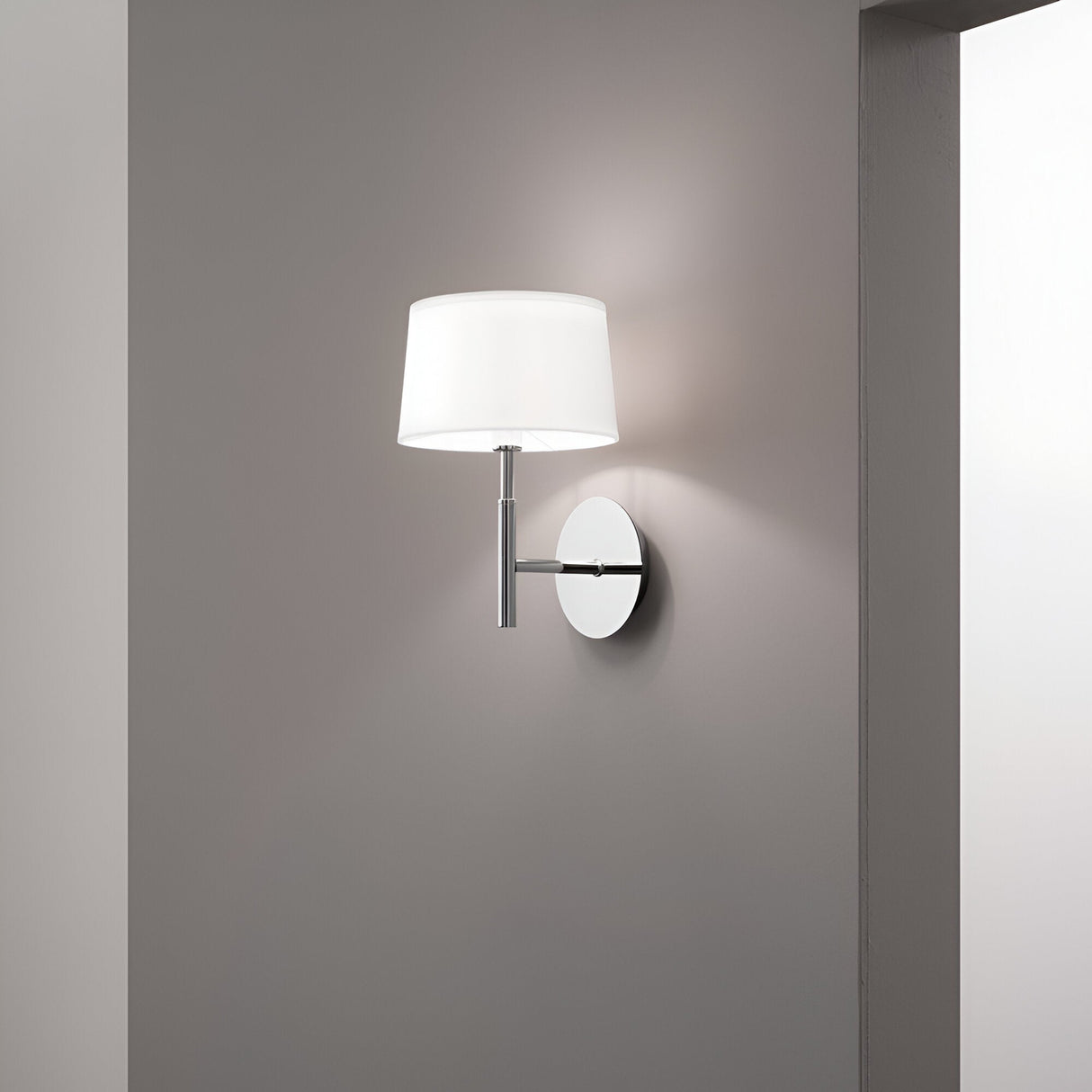 The Satin Wall Light - White, featuring a chrome base and white lampshade, is mounted on a neutral gray wall. When turned on, it emits a soft glow. The minimalist background with clean lines and muted colors perfectly exemplifies contemporary lighting design.