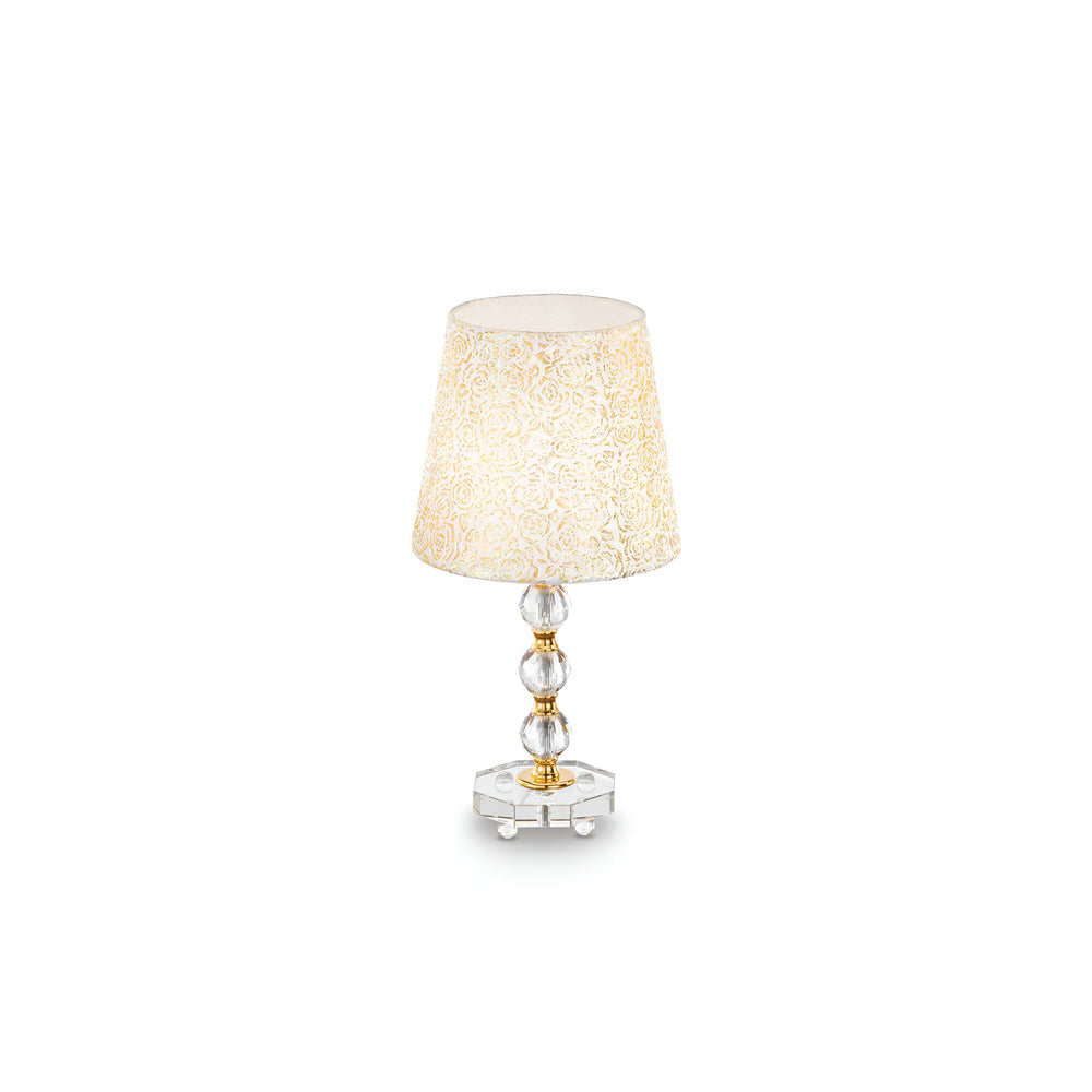The Princess Medium Crystal Table Lamp - Gold, with an elegant glass base featuring round, stacked crystals and a beige intricately patterned shade, gleams against a plain white background.