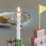 Lit white twist candle from the Twist Candles Pack of 12 in a green-patterned holder, set beside a patterned plate and a pink cake-shaped decor piece with a flag.