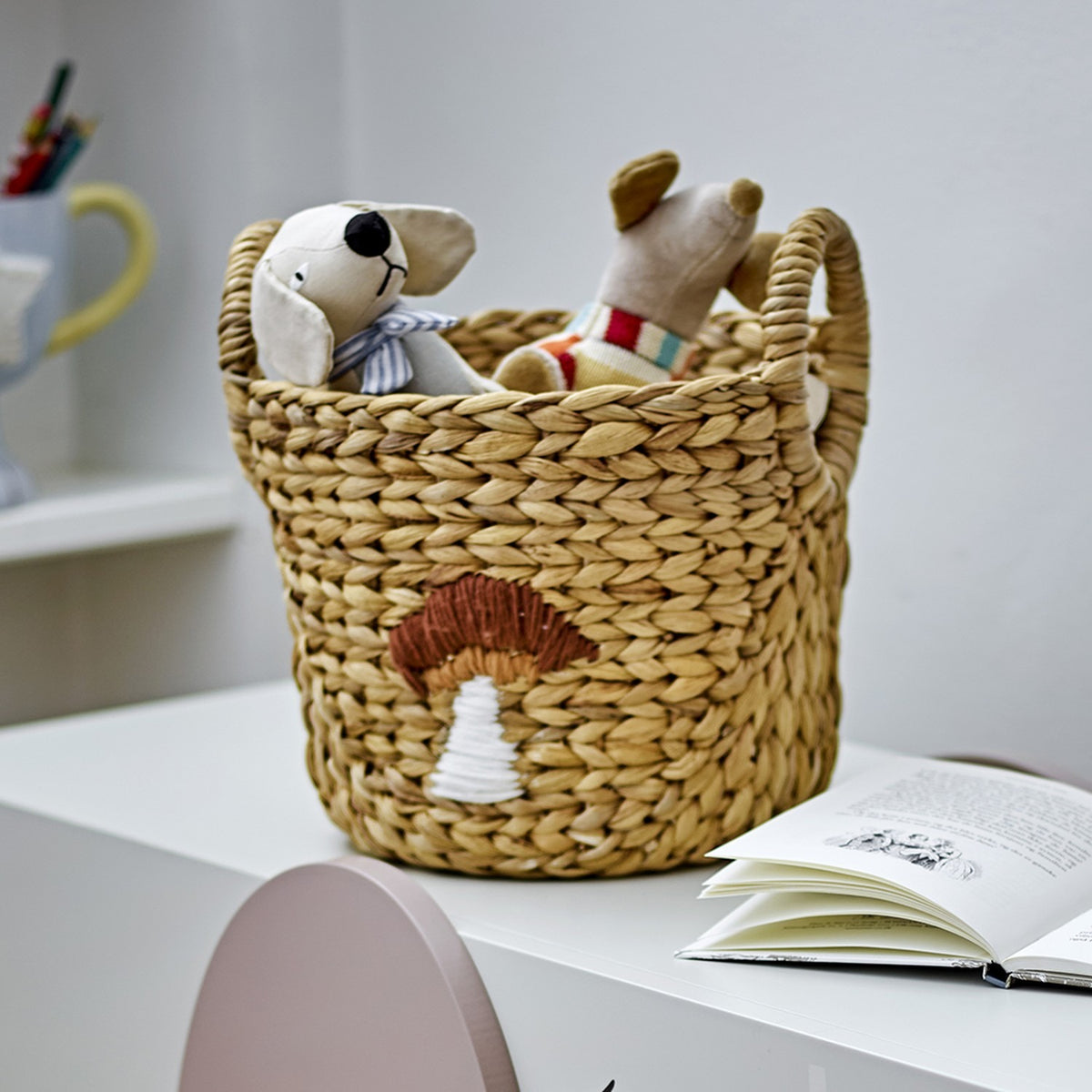 Agnes water hyacinth storage basket in beige with a stitched mushroom motif, holding soft toy mice, shown on a desk beside an open book.