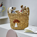Agnes water hyacinth storage basket in beige with a stitched mushroom motif, holding soft toy mice, shown on a desk beside an open book.