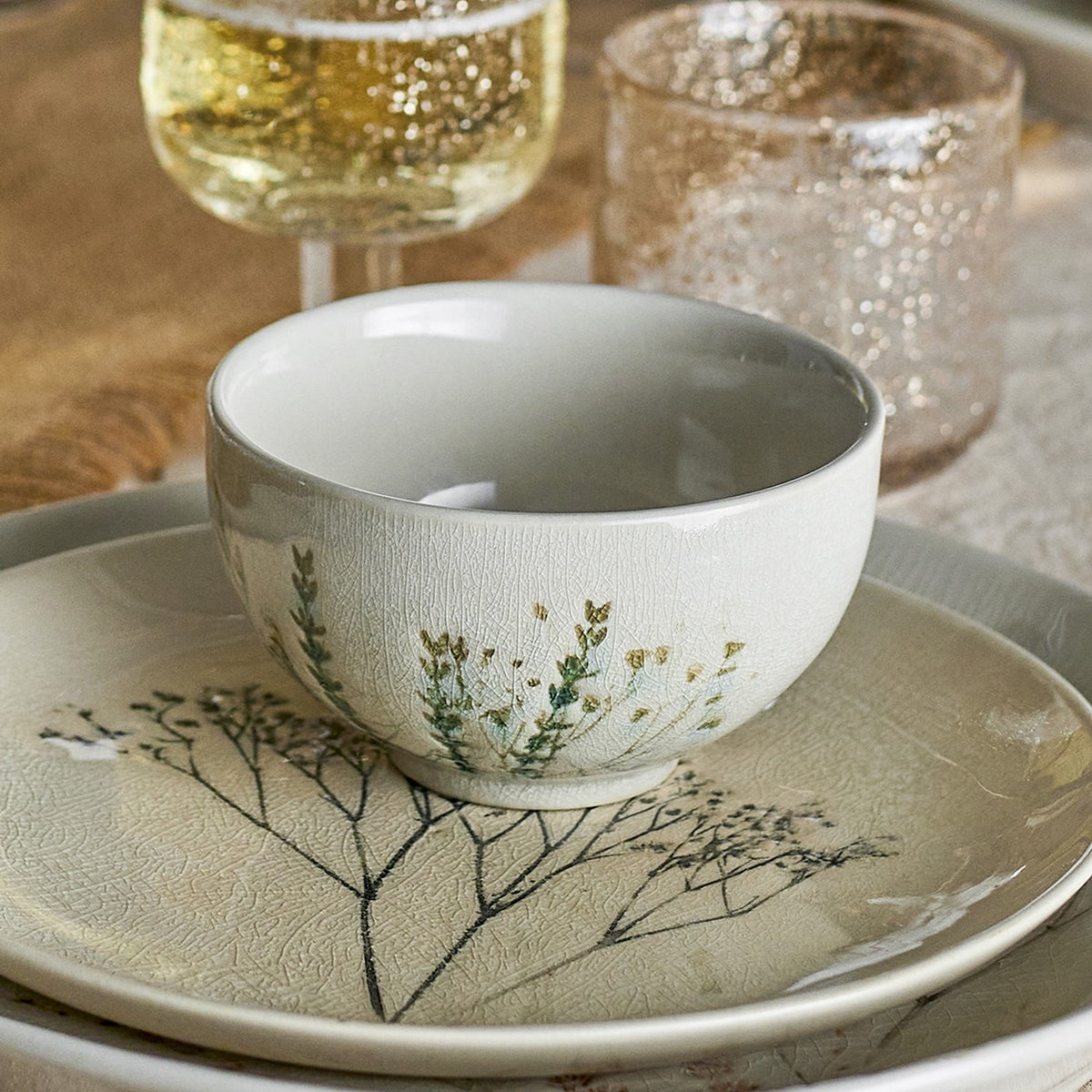 Cream crackle stoneware bowl with botanical grass motif resting on a matching patterned plate, part of the Bea 12-piece dinner set, shown with glassware in a table setting.